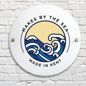 May include: White circular sign with the text "MAKES BY THE SEA" and "MADE IN KENT" in black. The sign features a blue and white wave design with a mountain and a yellow background.