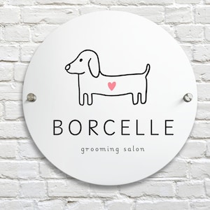 May include: A round white sign with the text "BORCELLE grooming salon". The sign features a black line drawing of a dachshund with a pink heart on its side. The sign is mounted on a white brick wall.