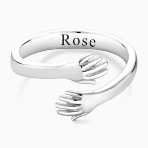 Puede incluir: Anillo de plata con dos manos entrelazadas en un abrazo. El anillo está grabado con el nombre "Rose".