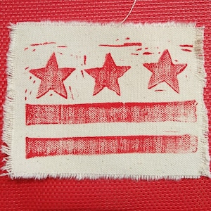 Puede incluir: Un parche de tela blanca con una impresión roja de tres estrellas y dos rayas horizontales. La tela tiene un borde deshilachado.