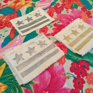 May include: Three fabric patches with a design of the flag of Washington, D.C. The patches are on a floral fabric background. The patches are in different shades of gray and gold.