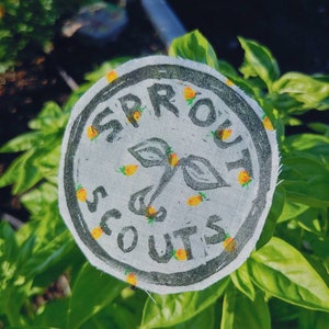 May include: A white fabric patch with black text that reads "Sprout Scouts" and a small green sprout with yellow fruit. The patch is surrounded by a black circle and has small yellow fruit scattered around the edge.