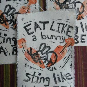 May include: A white fabric patch with black text that reads "EAT LIKE A BUNNY STING LIKE A BEE". The patch features a black and orange illustration of a bee.