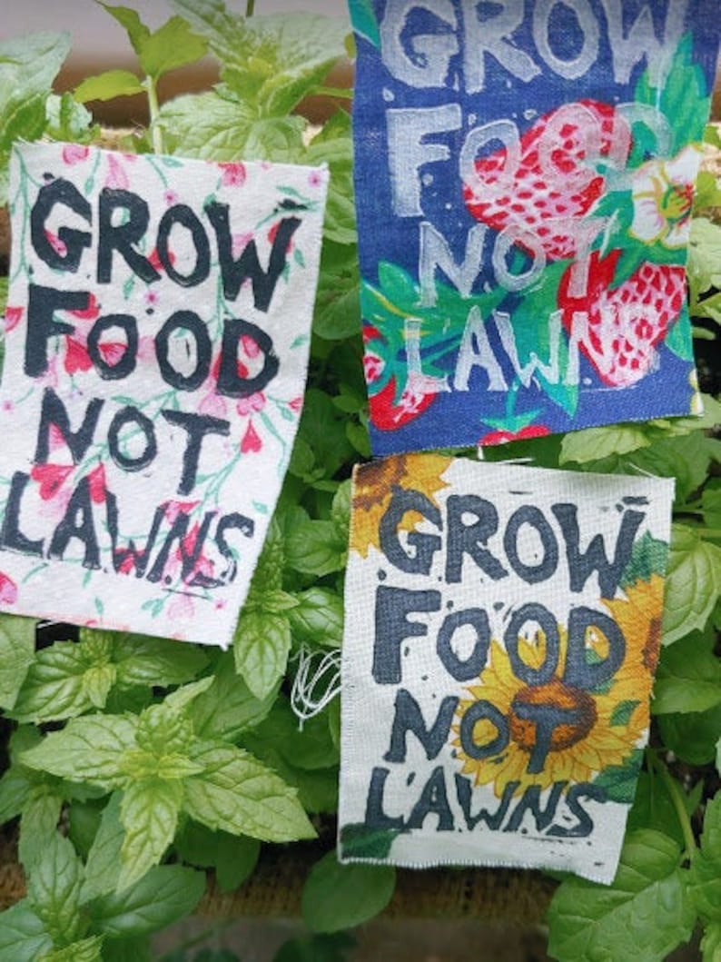May include: Three fabric patches with the text "Grow Food Not Lawns" in black lettering on a white background. The patches are decorated with different floral patterns. The first patch has pink and white flowers, the second patch has blue and white flowers, and the third patch has yellow and white flowers.