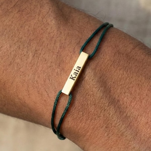 Peut inclure: Un bracelet avec une barre dorée gravée du nom "Kaia". La barre est reliée à un bracelet en cordon vert foncé. Le bracelet est porté sur le bras d'une personne.