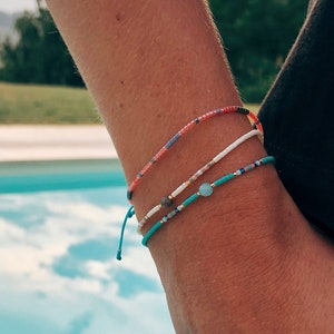 Può includere: Tre braccialetti colorati con perline al polso di una persona. I braccialetti sono fatti di piccole perline di vari colori, tra cui blu, rosso, bianco e verde. I braccialetti sono legati con un nodo alla fine.