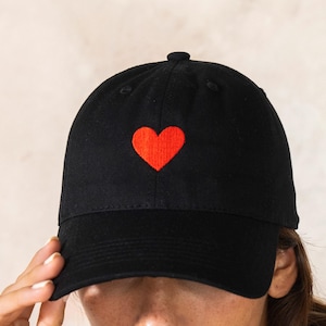 Puede incluir: Gorra de béisbol negra con un corazón rojo bordado en la parte delantera. La gorra tiene una visera curva y una correa ajustable. Un accesorio sencillo y elegante.