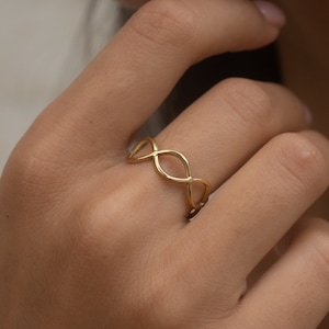 May include: A gold-colored ring featuring an infinity symbol design. The ring is worn on a finger, showcasing its delicate and intricate pattern. The ring's design consists of interconnected loops, creating a continuous, flowing shape. The ring is a piece of jewelry.
