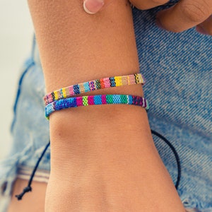Puede incluir: Dos pulseras tejidas coloridas con un patrón de rayas se muestran en una muñeca. Las pulseras presentan una variedad de colores, incluyendo azul, rosa, amarillo y verde. Las pulseras están aseguradas con una cuerda negra.