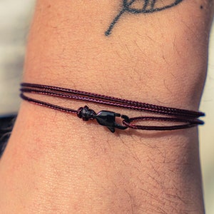 Puede incluir: Un brazalete de cordón rojo oscuro con un cierre negro. El brazalete se lleva en la muñeca.