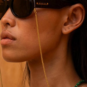 Op de afbeelding: Een vrouw draagt een zwarte ovale zonnebril met een gouden ketting die aan de pootjes is bevestigd. De ketting is ook bevestigd aan een groene kralenketting.