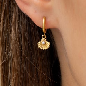 Puede incluir: Pendiente de aro dorado con un dije de concha. El dije es un diseño detallado de concha de vieira con un pequeño acento redondo blanco similar a una perla en el centro. El pendiente se muestra en una oreja.