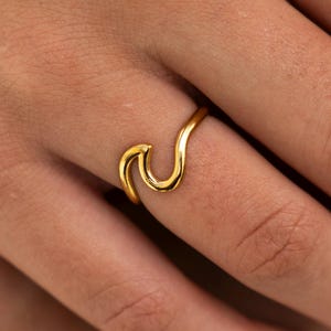 Könnte beinhalten: Ein goldener Wellenring an einem Finger. Der Ring ist ein schlichtes Design mit einer geschwungenen Wellenform.