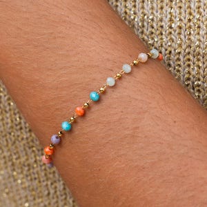 Op de afbeelding: Een kralenarmband met een gouden ketting en kleurrijke kralen. De kralen zijn in tinten oranje, turquoise, paars en wit. De armband wordt om een arm gedragen, tegen een goudkleurige achtergrond.