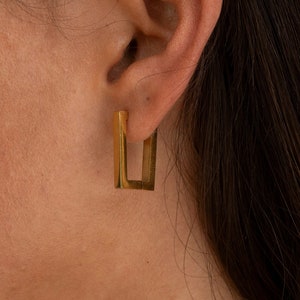 Peut inclure: Boucles d'oreilles carrées en or avec un design géométrique simple. Les boucles d'oreilles sont présentées sur l'oreille d'une personne.