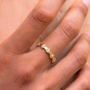 Op de afbeelding: Een gouden ring met een delicaat ontwerp van kleine bloemen. De ring wordt gedragen aan de vinger van een hand.