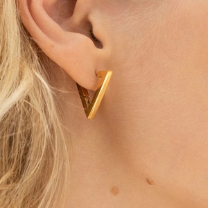 May include: Close-up of a gold-colored, triangular earring. The earring is a simple, geometric design, with a solid, polished surface. The earring is worn on a person's ear, with the focus on the jewelry.