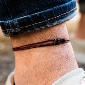 Könnte beinhalten: Nahaufnahme eines Knöchels, der ein dünnes, dunkelrotes Armband aus Schnur mit einem kleinen schwarzen Perlenakzent trägt. Das Armband wird über einem Paar weißen Wildlederschuhen getragen.