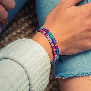 Könnte beinhalten: Eine Person trägt blaue Jeans und einen grauen Pullover mit zwei bunten Armbändern am Handgelenk.