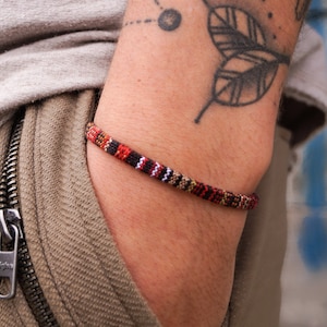 May include: A close-up of a person's arm wearing a multi-colored woven bracelet. The bracelet is made of red, black, white, and brown threads. The person has a tattoo of a leaf on their arm.