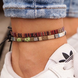 May include: Two colorful woven ankle bracelets with black string ties. The bracelets are worn on a person's ankle above white sneakers.