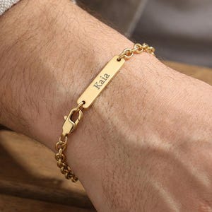 Könnte beinhalten: Ein goldfarbenes Metallkettenarmband mit einer rechteckigen Identifikationsplatte, in die der Name Kaia eingraviert ist. Das Armband wird mit einem Karabinerverschluss gesichert und zeichnet sich durch ein poliertes Finish und ein klassisches Gliederkettendesign aus.