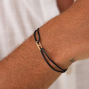 Könnte beinhalten: Ein schwarzes Kordelarmband mit einem goldfarbenen rechteckigen Verschluss. Das Armband wird am Handgelenk einer Person getragen, vor dem Hintergrund eines weißen Hemdes mit Knöpfen. Das Design des Armbands ist schlicht und elegant.