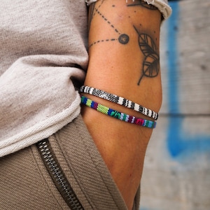 Könnte beinhalten: Ein Arm mit einem schwarz-weißen Blatttattoo, der zwei Armbänder trägt. Das obere Armband ist schwarz-weiß mit einem geometrischen Muster. Das untere Armband ist mehrfarbig mit einem gewebten Muster.