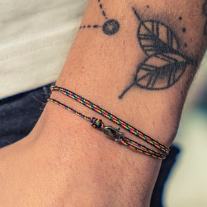 Puede incluir: Un brazalete trenzado de colores con un cierre plateado. El brazalete se lleva en la muñeca.