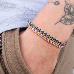 Könnte beinhalten: Eine Person trägt ein braunes Hemd mit Reißverschluss und ein mehrfarbiges geflochtenes Armband am Handgelenk.