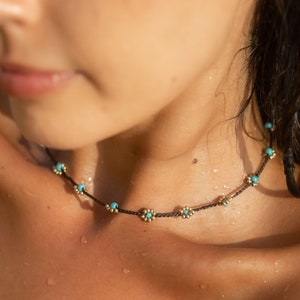 Può includere: Primo piano del collo di una donna con una delicata collana choker nera con piccole perle di fiori turchesi e dorate.
