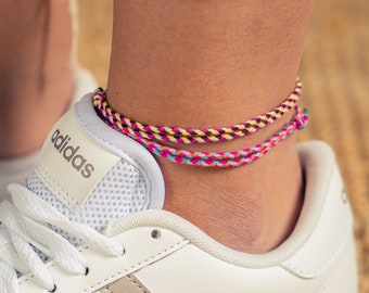 May include: White leather sneakers with three silver stripes and the word "adidas" on the side. The sneakers are worn by a person wearing blue jeans and a colorful ankle bracelet.