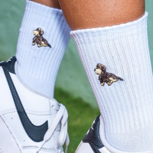 May include: A pair of white ribbed crew socks featuring a small embroidered brown and black dog graphic on the outer side of each sock, paired with white leather sneakers that have a black swoosh logo on the side and white laces.