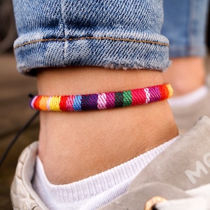 May include: A colorful woven ankle bracelet with a black string tie. The bracelet is made of multiple strands of yarn in various colors, including red, orange, yellow, green, blue, purple, and pink.