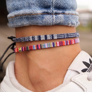 May include: Two colorful woven anklets on a person's ankle. The top anklet is navy blue and white, and the bottom anklet is multi-colored with red, pink, yellow, and blue.