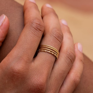 Puede incluir: Primer plano de una mano con cuatro anillos apilables de oro. Los anillos son finos y delicados, con una superficie texturizada.