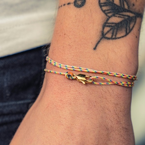 Puede incluir: Primer plano de una muñeca adornada con dos pulseras de cuerda coloridas. Las pulseras, con un cierre dorado, están hechas de hilos entrelazados en tonos amarillos, azules, verdes y rojos. El brazo tiene un tatuaje de hojas.