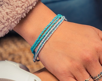 May include: A close-up of a person's wrist wearing a blue and white braided bracelet. The person is also wearing two silver rings on their fingers, one with a leaf design and the other with a small gemstone.