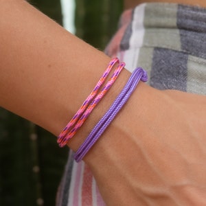Può includere: Due braccialetti su un polso. Uno è un braccialetto in corda viola, l'altro è un braccialetto in corda rosa e arancione.