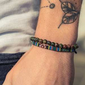 Op de afbeelding: Een close-up van iemands pols met twee armbanden. De bovenste armband is gemaakt van donkergroene en bruine kralen. De onderste armband is een geweven band met rode, gele, blauwe en zwarte strepen.