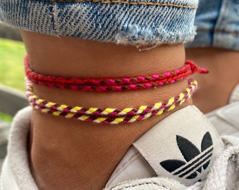 May include: Two colourful braided anklets on a person's ankle. The top anklet is red, pink, and brown. The bottom anklet is yellow, brown, and white.