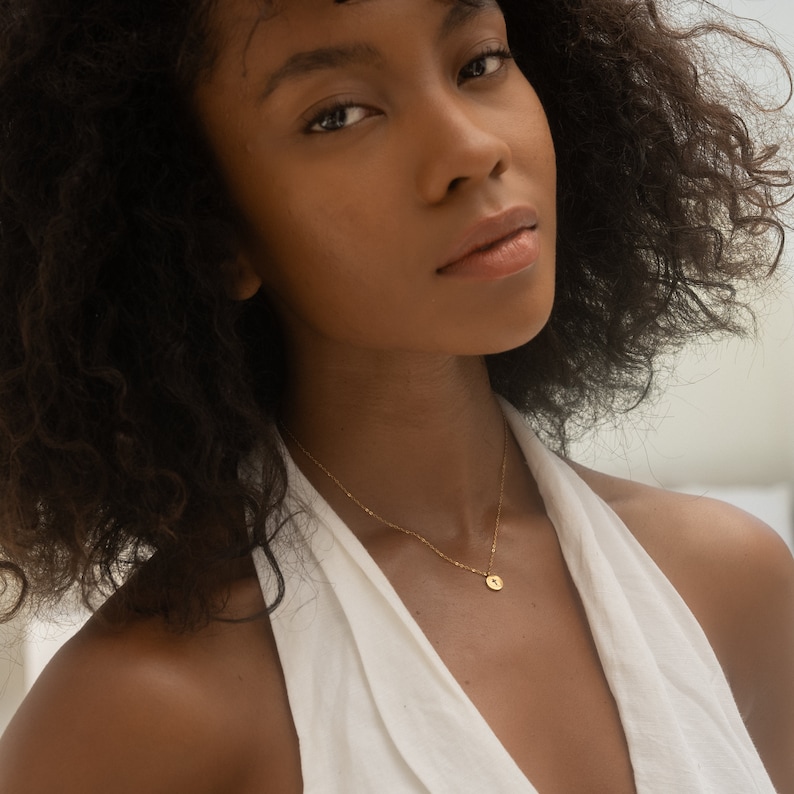 May include: A woman with curly dark brown hair wearing a white halter top and a gold chain necklace with a small round pendant.
