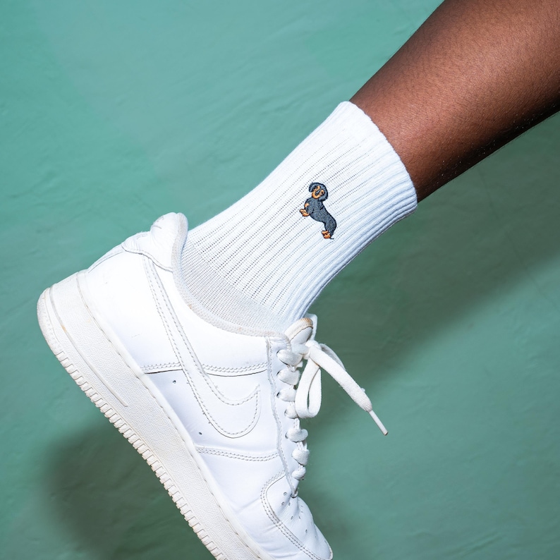 May include: White athletic socks with a small embroidered dachshund on the side. The socks are being worn with white trainers.