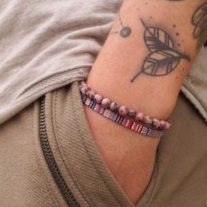 Könnte beinhalten: Eine Person trägt ein hellbraunes Hemd und eine Hose mit Reißverschlussdetail. Sie trägt zwei Armbänder am linken Handgelenk, eines ist ein Perlenarmband mit rosa und weißen Perlen, das andere ist ein gewebtes Armband mit roten, blauen und weißen Streifen.