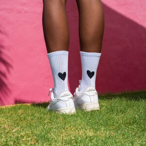 May include: A pair of white ribbed crew socks featuring a small black heart graphic on the outer side of each ankle, paired with white low-top athletic sneakers. The socks are mid-calf length and made of a knit fabric with a visible vertical texture.