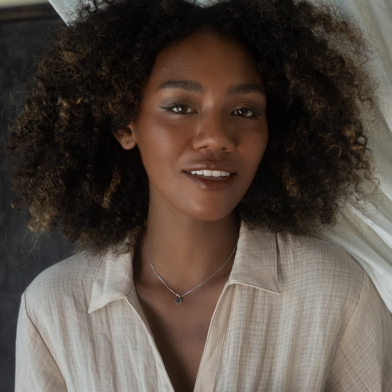 May include: A woman with curly brown hair wearing a light brown shirt with a silver necklace with a small pendant.