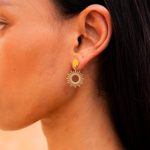 Könnte beinhalten: Goldfarbene Ohrringe mit Sonnenstrahlen-Design. Die Ohrringe bestehen aus kleinen Perlen, die in einem Kreis angeordnet sind, mit Strahlen, die nach außen verlaufen. Die Ohrringe hängen an einem kleinen goldenen Kreis.