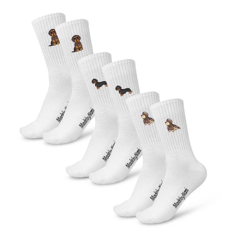 May include: Set of six white crew socks with embroidered dachshund designs. The socks feature the text "MadebyNomi" on the bottom of each sock.