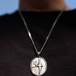May include: A silver chain necklace with an oval pendant featuring a detailed, engraved image of a bird in flight.
