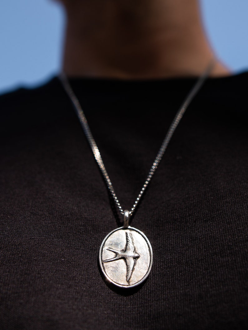 May include: A silver chain necklace with an oval pendant featuring a detailed, engraved image of a bird in flight.
